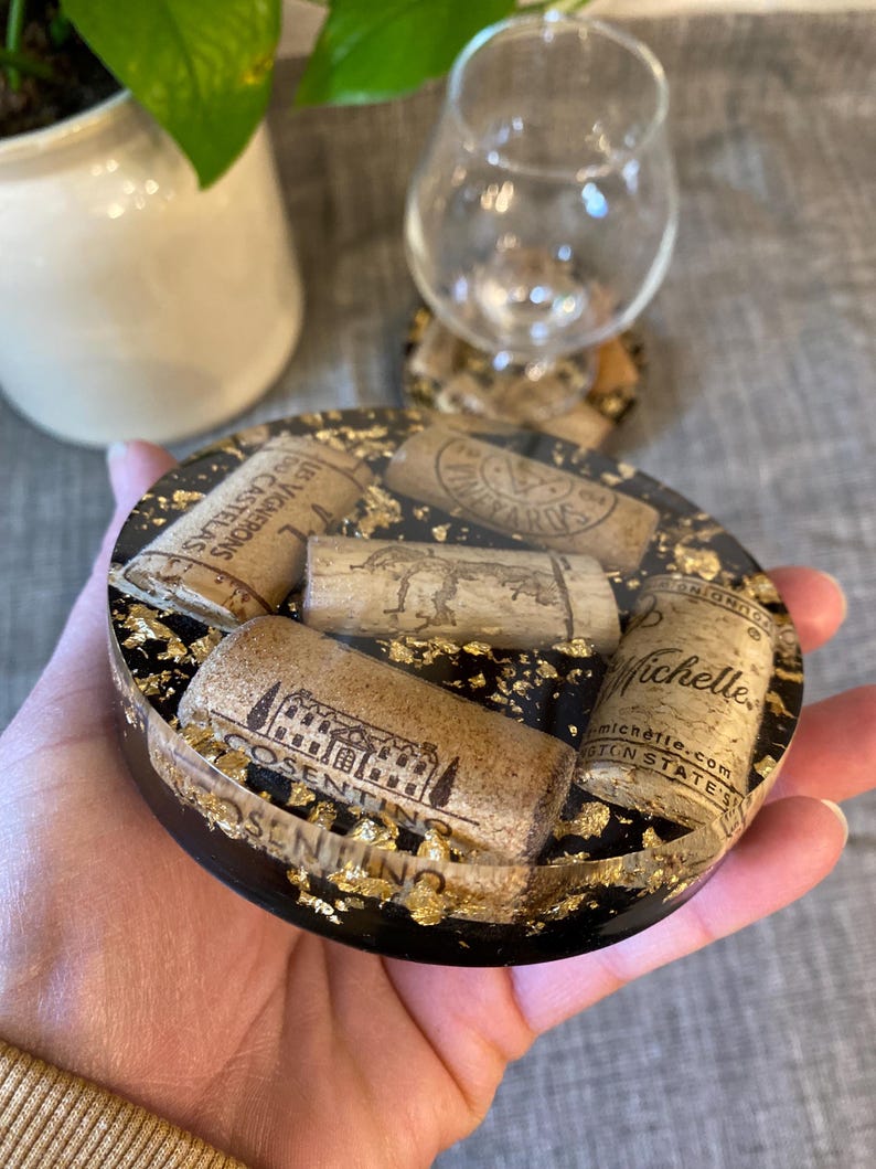 May include: A round black resin coaster with gold leaf accents, embedded with several wine corks. The corks have various winery names and logos printed on them. A wine glass sits nearby.