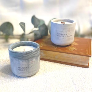 Può includere: Due candele di cera di soia in contenitori di cemento. Una candela è etichettata "Banana Coffee Cake", l'altra "Hansel & Gretel". Le candele sono bianche con stoppini di legno. I contenitori sono grigi e bianchi, su un libro marrone.