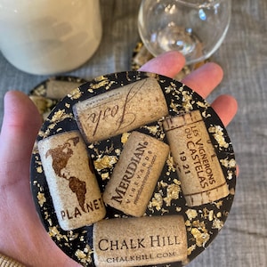 May include: Round black coaster with gold flecks, embedded with wine corks. Corks include the words "Josh", "Meridian Vineyards", "Planet", "Chalk Hill", and "Les Vignerons Du Castelas 1921".