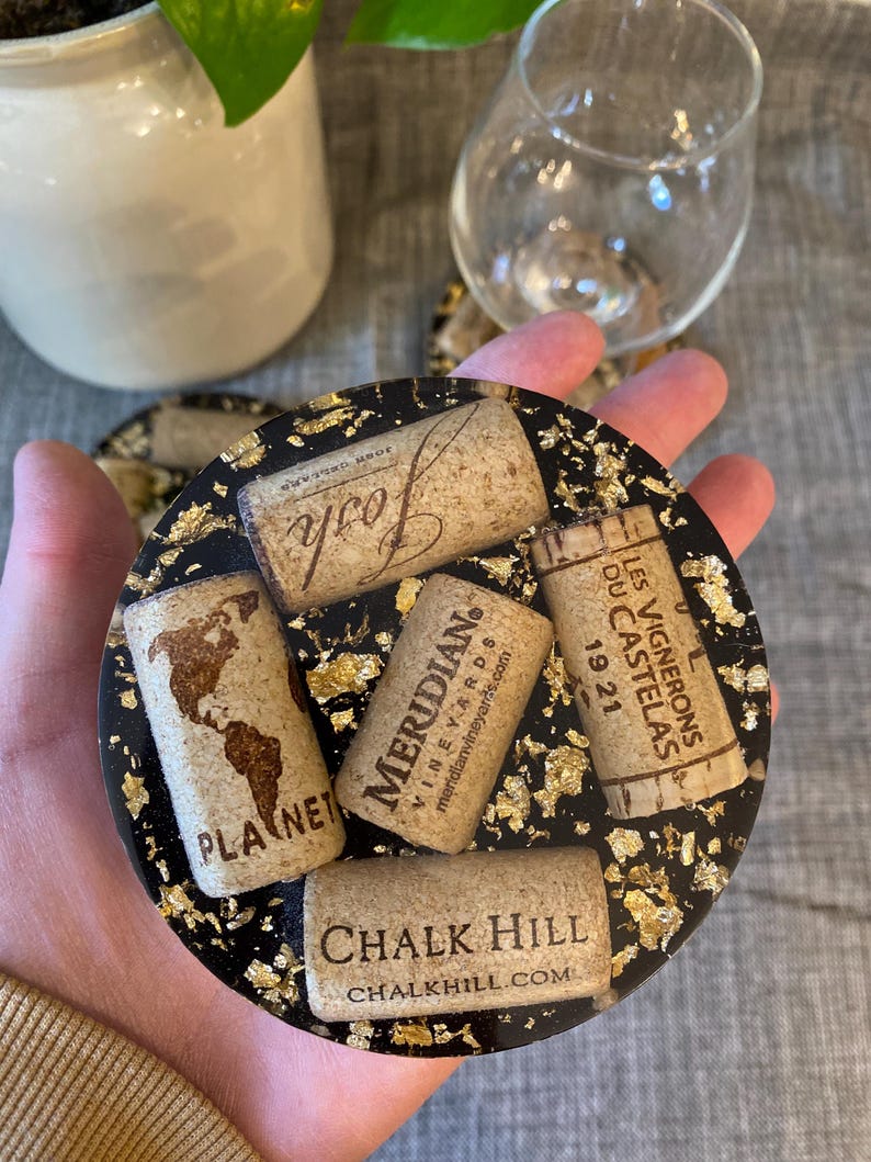 May include: Round black coaster with gold flecks, embedded with wine corks. Corks include the words "Josh", "Meridian Vineyards", "Planet", "Chalk Hill", and "Les Vignerons Du Castelas 1921".
