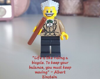 May include: A yellow Lego figurine of Albert Einstein holding a brown stick. The figurine is wearing a brown jacket, a white shirt, a bow tie, and black pants. The text "Life is like riding a bicycle. To keep your balance, you must keep moving" - Albert Einstein is written below the figurine.