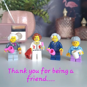 Könnte beinhalten: Vier Lego-Figuren, die jeweils eine Figur darstellen, stehen in einer Reihe. Die Figuren tragen verschiedene Outfits und halten Accessoires wie eine rosa Gießkanne und eine rote Handtasche. Der Text "Thank you for being a friend...." ist rosa geschrieben.