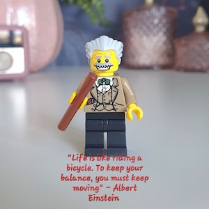 Peut inclure: Une figurine Lego jaune d'Albert Einstein tenant un bâton brun. La figurine porte une veste brune, une chemise blanche, un nœud papillon et un pantalon noir. Le texte "Life is like riding a bicycle. To keep your balance, you must keep moving" - Albert Einstein est écrit sous la figurine.