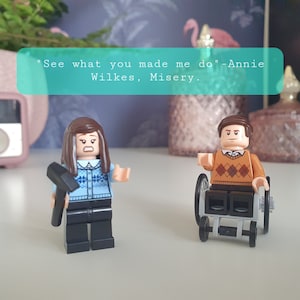 Peut inclure: Deux figurines Lego, une femme aux cheveux bruns tenant un marteau et portant un pull bleu à motifs blancs et bleus, et un homme aux cheveux bruns portant un pull marron à motifs losanges blancs et marrons, assis dans un fauteuil roulant. La citation "See what you made me do" - Annie Wilkes, Misery est écrite dans un encadré turquoise.