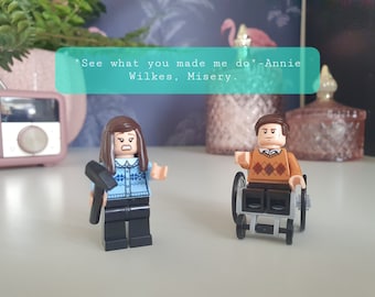 May include: Two Lego minifigures, one female with brown hair holding a hammer and wearing a blue sweater with a white and blue pattern, and one male with brown hair wearing a brown sweater with a white and brown diamond pattern, sitting in a wheelchair. The quote "See what you made me do" - Annie Wilkes, Misery is written in a teal box.