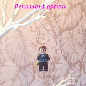 May include: A Lego figure ornament with a silver hook. The figure is wearing a black jacket, blue shirt, and black pants. The figure is holding a gold-colored object. The text "Ornament option" is in pink.