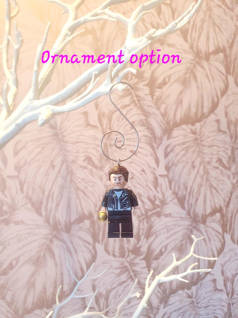 May include: A Lego figure ornament with a silver hook. The figure is wearing a black jacket, blue shirt, and black pants. The figure is holding a gold-colored object. The text "Ornament option" is in pink.