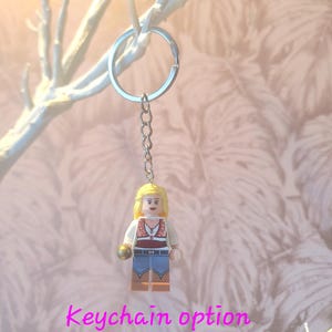 May include: A keychain featuring a Lego figurine of a woman with blonde hair, wearing a white shirt, red vest, and blue jeans. The keychain has a silver ring and chain. The text "Keychain option" is displayed in pink.