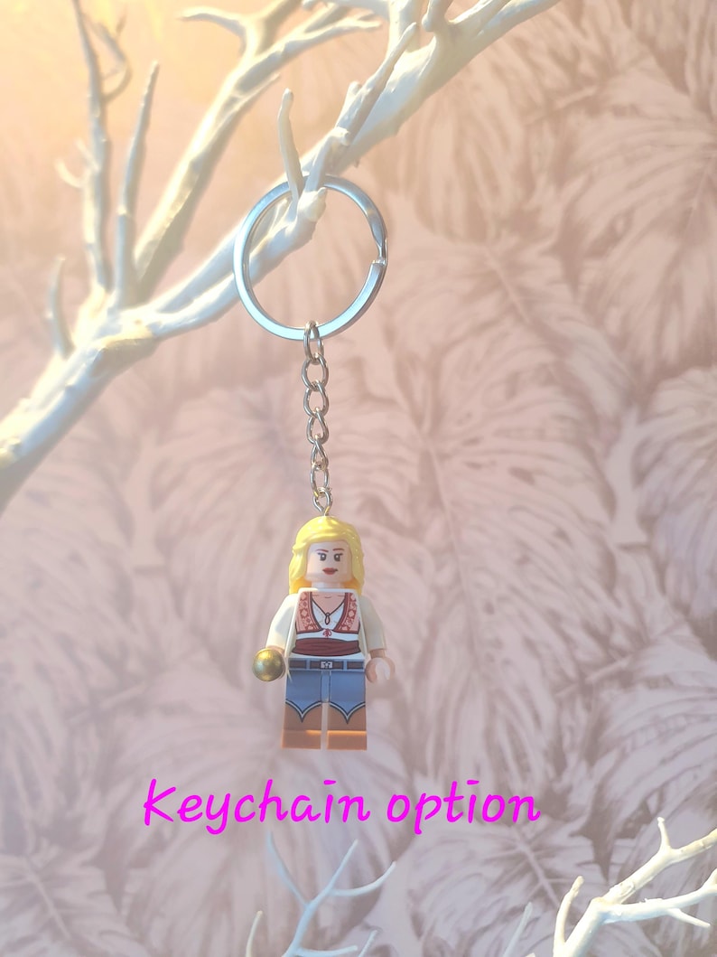 May include: A keychain featuring a Lego figurine of a woman with blonde hair, wearing a white shirt, red vest, and blue jeans. The keychain has a silver ring and chain. The text "Keychain option" is displayed in pink.