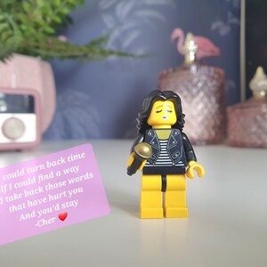 Może przedstawiać: Żółta figurka Lego kobiety z czarnymi włosami i czarną skórzaną kurtką trzymająca złoty mikrofon. Figurka stoi na białym tle. Różowa karteczka z tekstem piosenki "If I Could Turn Back Time" Cher znajduje się na pierwszym planie.