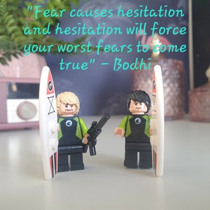 Puede incluir: Dos minifiguras Lego con trajes de neopreno verde y negro, sosteniendo tablas de surf. Las minifiguras están de pie sobre una superficie blanca. La cita "El miedo causa la vacilación y la vacilación obligará a tus peores miedos a hacerse realidad" - Bodhi es visible en el fondo.