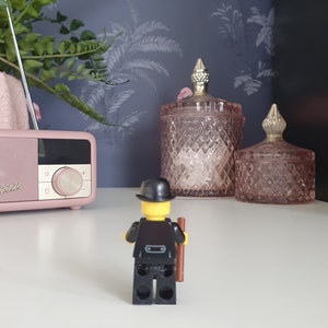 Puede incluir: Una figura Lego negra que lleva un sombrero de copa negro y sostiene un palo marr&oacute;n. La figura est&aacute; de pie sobre una superficie blanca frente a dos tarros de vidrio rosa con tapas doradas.