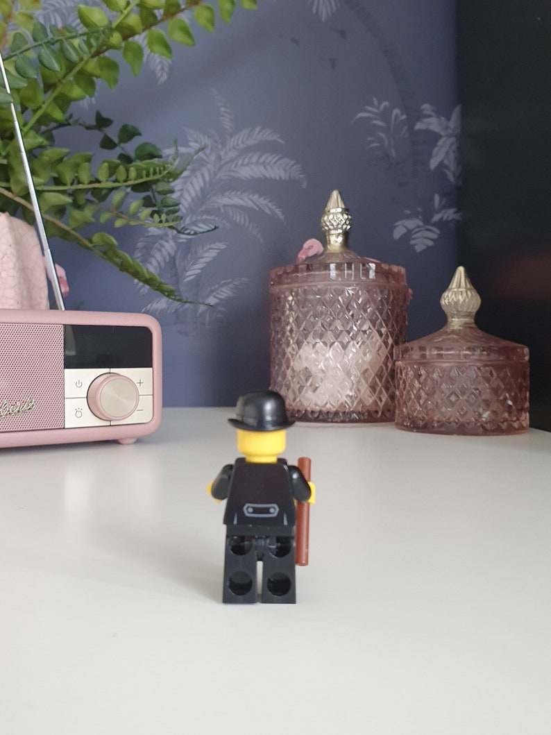 Puede incluir: Una figura Lego negra que lleva un sombrero de copa negro y sostiene un palo marr&oacute;n. La figura est&aacute; de pie sobre una superficie blanca frente a dos tarros de vidrio rosa con tapas doradas.