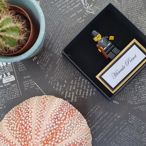 Puede incluir: Una caja de regalo negra y dorada con una figura de Lego de H&eacute;rcules Poirot, un detective ficticio. La caja tiene una tarjeta blanca con el texto "Hercule Poirot" en letras negras.