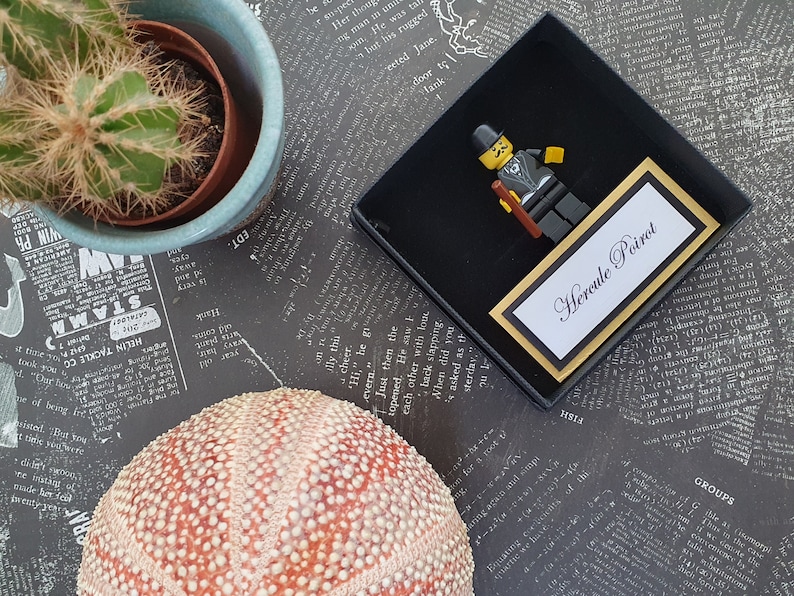 Puede incluir: Una caja de regalo negra y dorada con una figura de Lego de H&eacute;rcules Poirot, un detective ficticio. La caja tiene una tarjeta blanca con el texto "Hercule Poirot" en letras negras.