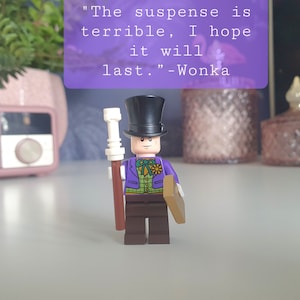 Puede incluir: Una minifigura LEGO de Willy Wonka de color morado y verde, con un sombrero de copa negro y un bastón marrón y blanco. La minifigura está de pie sobre una superficie blanca. La cita "La suspense es terrible, espero que dure." - Wonka es visible en el fondo.