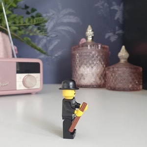 Puede incluir: Una minifigura Lego negra y amarilla con un sombrero negro y un palo marr&oacute;n. La minifigura est&aacute; de pie sobre una superficie blanca.
