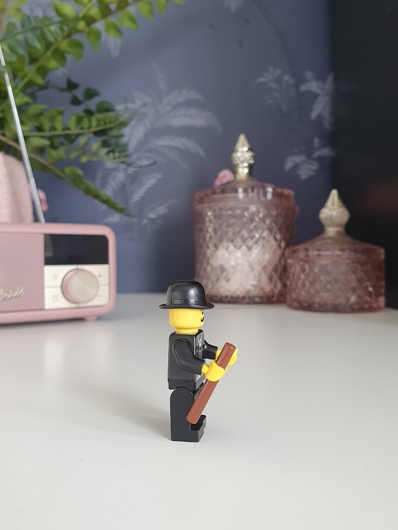 Puede incluir: Una minifigura Lego negra y amarilla con un sombrero negro y un palo marr&oacute;n. La minifigura est&aacute; de pie sobre una superficie blanca.