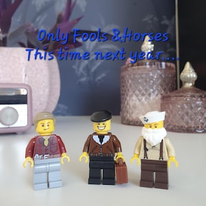 May include: Three Lego minifigures, one wearing a brown jacket, one wearing a red shirt, and one wearing a white sailor hat and a white beard. The text "Only Fools & Horses This time next year..." is written above the figures.