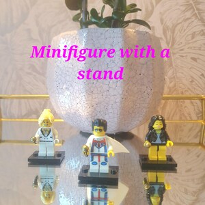 May include: Three Lego minifigures on black stands, reflected on a mirrored surface. The figures include a blonde woman in a white suit, a superhero, and a woman in a black jacket. A white and pink cracked ceramic pot with a plant is in the background.