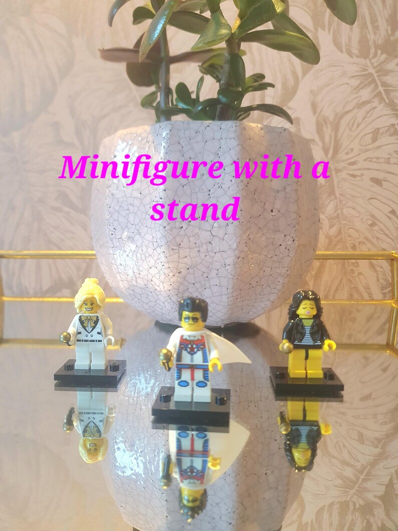 May include: Three Lego minifigures on black stands, reflected on a mirrored surface. The figures include a blonde woman in a white suit, a superhero, and a woman in a black jacket. A white and pink cracked ceramic pot with a plant is in the background.