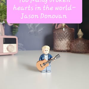 May include: A Lego figurine of a person wearing a blue denim jacket and light blue jeans, holding a brown acoustic guitar. The figurine is standing on a white surface. The text "Too Many broken hearts in the world - Jason Donovan" is written in pink on a pink background.