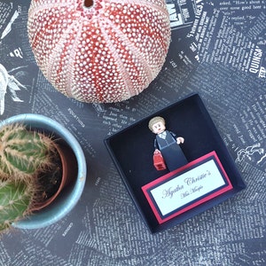 May include: A Lego figure of Miss Marple from Agatha Christie's novels, holding a red purse, inside a black box with a nameplate. A sea urchin and a cactus in a blue pot are also in the image.
