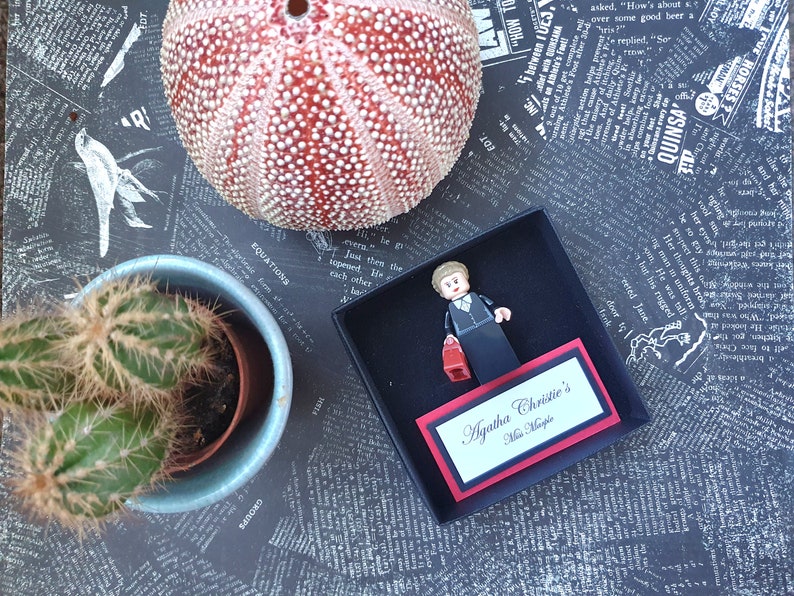 May include: A Lego figure of Miss Marple from Agatha Christie's novels, holding a red purse, inside a black box with a nameplate. A sea urchin and a cactus in a blue pot are also in the image.