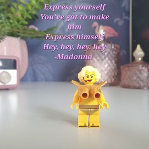 May include: A yellow Lego figurine of a woman with blonde hair wearing a gold-colored top and bottom. The figurine is holding a microphone and has a serious expression on her face. The background is a blurry image of a pink radio and a pink and gold decorative box. The text "Express yourself You've got to make him Express himself Hey, hey, hey, hey -Madonna" is written in pink on a blue background.