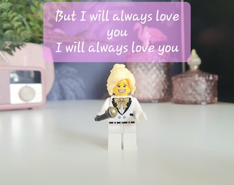 Könnte beinhalten: Eine Lego-Figur einer Frau mit blonden Haaren, die einen weißen Anzug mit schwarzer Fliege trägt und ein Mikrofon hält. Der Hintergrund ist verschwommen und zeigt ein rosa Radio und eine rosa-weiße Dekobox. Der Text "But I will always love you I will always love you" ist in einem rosa Kasten über der Figur geschrieben.