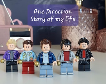 May include: Five Lego figures of a boy band, each with distinct hair and clothing. The figures are arranged in a line. The text "One Direction Story of my life" is displayed on a purple banner above the figures.
