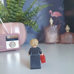 May include: A Lego figurine of a person with blonde hair in a bun, wearing a black outfit and holding a red bag. The figurine is facing away from the camera. A pink radio and a potted plant are in the background.