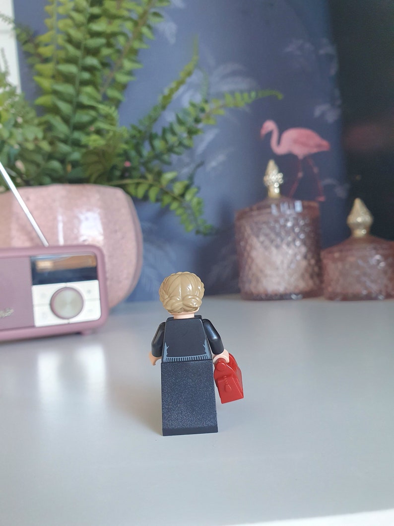 May include: A Lego figurine of a person with blonde hair in a bun, wearing a black outfit and holding a red bag. The figurine is facing away from the camera. A pink radio and a potted plant are in the background.