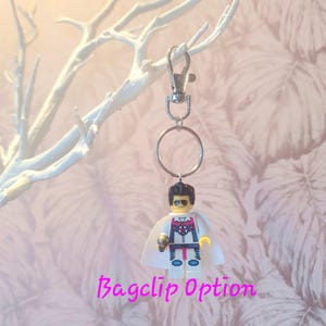 May include: A Lego minifigure keychain with a silver clasp and ring. The minifigure has a white cape, sunglasses, and a detailed costume. The text "Bagclip Option" is written in pink cursive below the keychain.