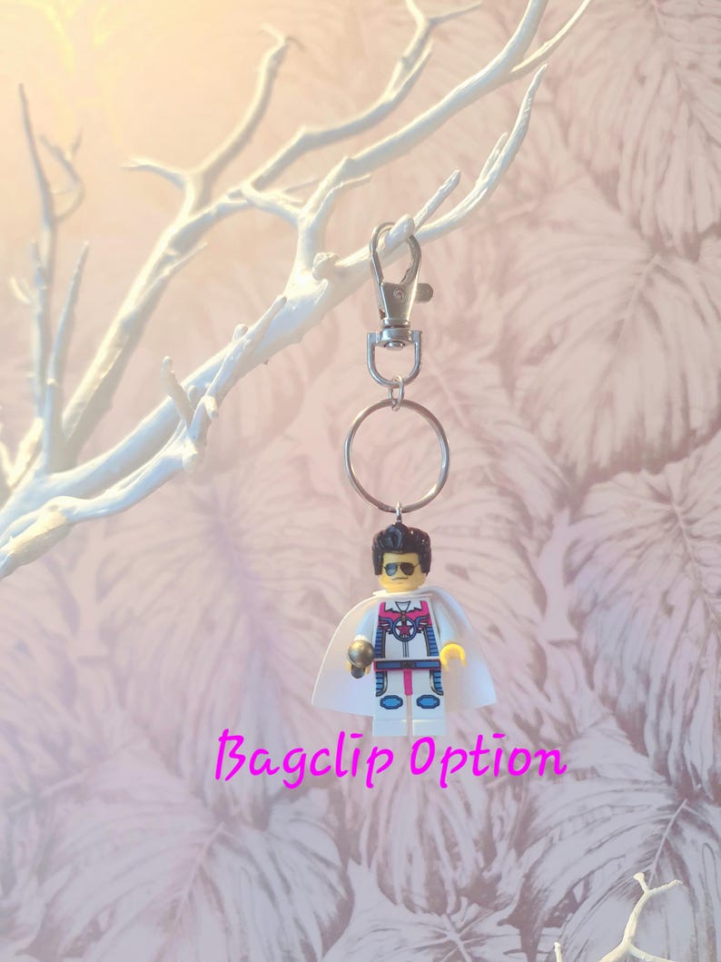 May include: A Lego minifigure keychain with a silver clasp and ring. The minifigure has a white cape, sunglasses, and a detailed costume. The text "Bagclip Option" is written in pink cursive below the keychain.