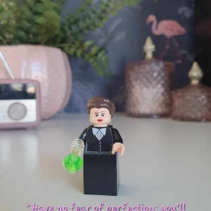 Pode incluir: Uma minifigura Lego preta de uma mulher com cabelo castanho, vestindo um vestido preto e segurando um copo de vidro verde. A citação "Não tenha medo da perfeição; você nunca a alcançará" está escrita abaixo da figura.