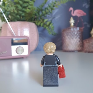 May include: A Lego figurine, viewed from the back, with blonde hair in a bun, wearing a black outfit and holding a red bag. The figurine is on a white surface, with a pink radio and a potted plant in the background.
