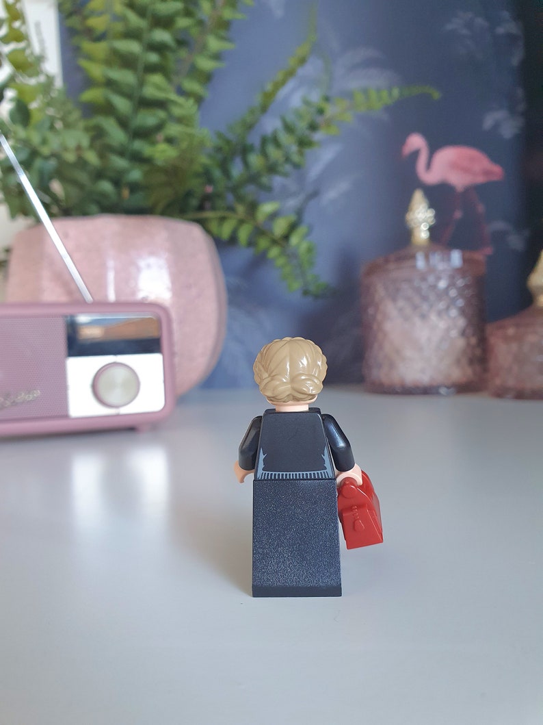 May include: A Lego figurine, viewed from the back, with blonde hair in a bun, wearing a black outfit and holding a red bag. The figurine is on a white surface, with a pink radio and a potted plant in the background.