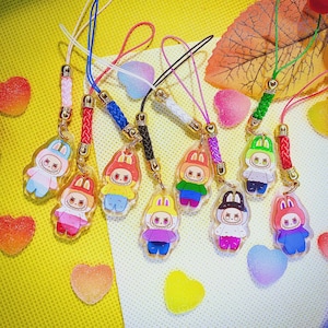 May include: A selection of acrylic charms with cartoon bunny characters in various outfits. Each charm is attached to a lanyard with a gold-coloured clasp. The background features heart-shaped sweets and a fallen leaf.