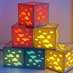 Può includere: Un set di sei blocchi Minecraft luminosi in vari colori, tra cui rosso, giallo, verde e blu. I blocchi sono impilati a forma di piramide, con il blocco più grande in basso e il blocco più piccolo in alto.