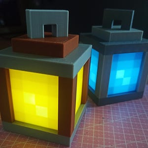 Pode incluir: Duas lanternas com tema Minecraft, uma com luz amarela e outra com luz azul. As lanternas são feitas de plástico cinza, marrom e azul e têm uma alça na parte superior.