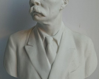 May include: A white bust sculpture of a man with a mustache and a suit. The sculpture is mounted on a blue and white circular base. The man has a stern expression.