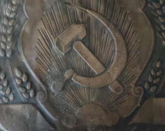 May include: A close-up of a bronze plaque with a hammer and sickle emblem, surrounded by wheat stalks and the inscription "USSR".