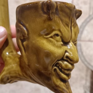 May include: A ceramic pipe with a devil face design. The pipe is a shade of brown with a glossy finish. The devil face has horns and a sinister grin, with detailed facial features.
