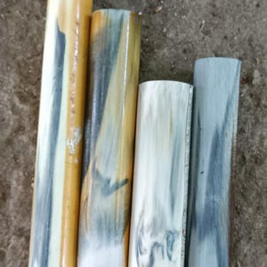 May include: Four cylindrical objects with a natural, layered appearance. The cylinders vary in colour, with shades of cream, tan, and grey. The objects appear to be made of a natural material, possibly bone or horn, and are arranged on a rough, grey surface.
