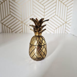 May include: A metallic gold-toned pineapple-shaped container with a removable lid. The object features a textured, cross-hatched diamond pattern on the body and a pointed, leafy top section. The surface has a weathered, distressed finish.
