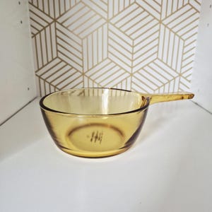 May include: A round, amber-coloured glass saucepan with a single handle extending from the side. The glass is translucent and smooth, featuring a clear, circular bowl shape with a flat bottom and a small, integrated handle for gripping.