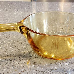 May include: An amber-coloured glass saucepan with a handle. The pan has a rounded bowl shape and a long, rectangular handle extending from the side. The glass has a slight golden hue, and the pan is on a speckled surface.