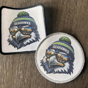 May include: Two patches featuring a stylized eagle wearing sunglasses and a Seahawks beanie. The eagle's sunglasses reflect a city skyline. One patch is square with a black border, the other is round with a silver border.