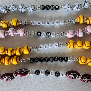 May include: Five colorful beaded bracelets with different sports-themed charms. The bracelets feature white and yellow baseballs, white and red baseballs, and brown footballs. The charms spell out "GAME DAY", "MAMA", "MAMA", "SOFTBALL", and "GAME DAY".